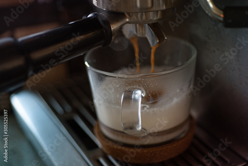 Espresso coffee is flowing out of the coffee maker to glass cappuccino cup with milk foam. Close up view.