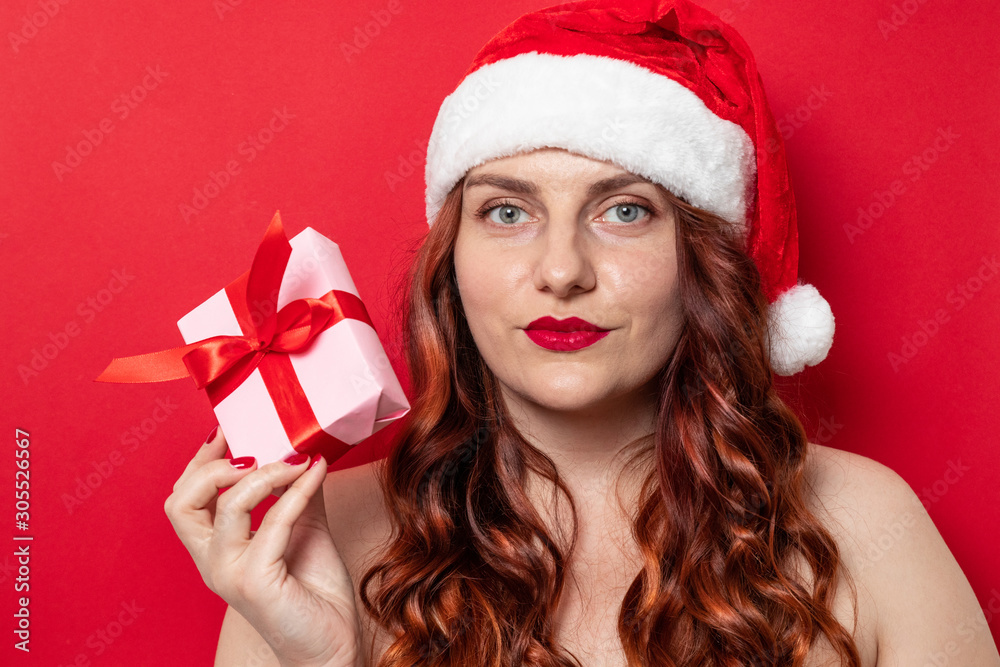 Portrait of a young curly girl with a red santa hat is holding a gift box with a red bow on a background of red wall. Portrait of a young curly girl with a red santa hat is holding a gift box with a red bow on a background of red wall.