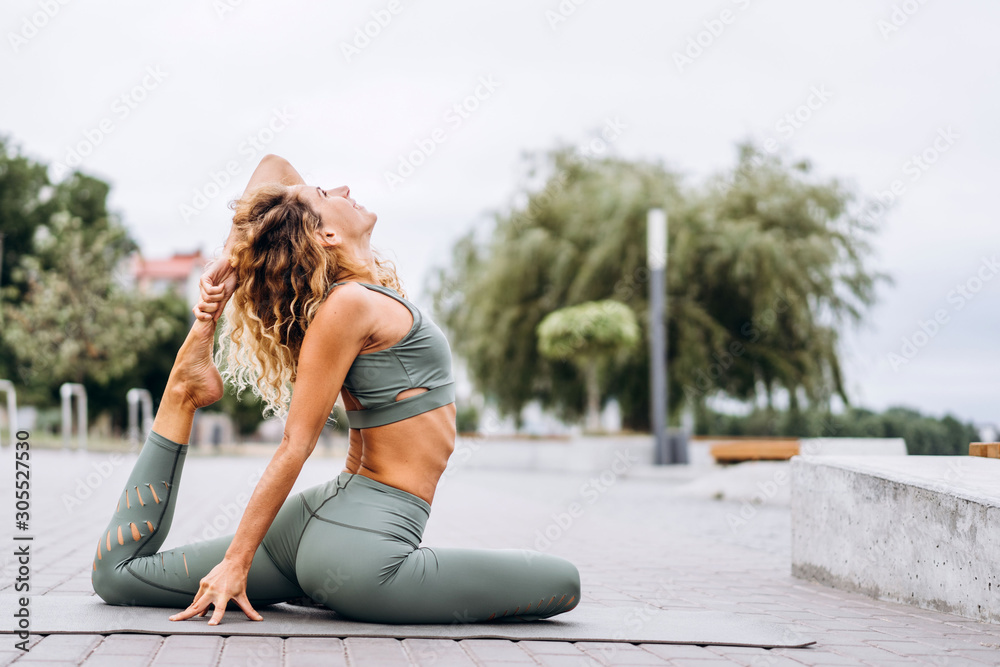 Sporty young woman with long hair in gray tracksuit doing stretching exercises on the street, on the background of trees. Active lifestyle, yoga concept