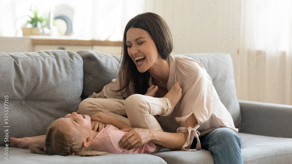 Cheerful mother tickling cute child daughter laughing playing on sofa