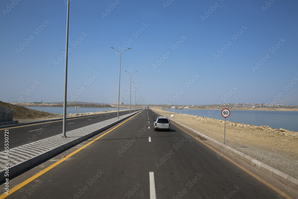 road under a blue sky with focus to the center white line . asphalt car road .wallpaper landscape .sea and lake road .light poles . Empty road white cloudy weather.