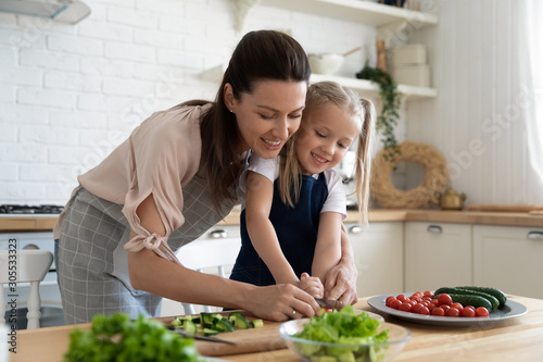 Happy mom teaching preschool daughter cutting salad in kitchen interior