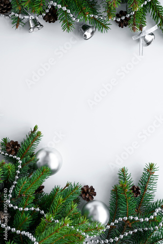 Christmas decor in silver and green on vertical postcard. Copy space surrounded by conifer branches, cones, baubles, bells and chain.