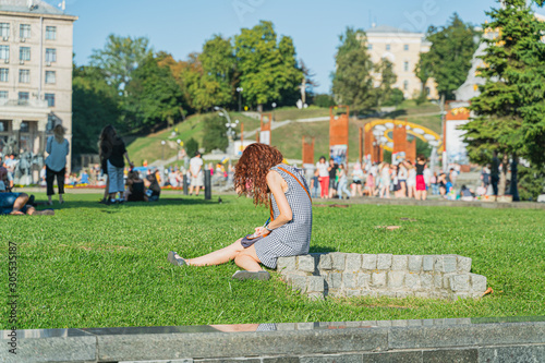 Casual student girl writing in the diary outdoors. Slim girl with curly red hair sitting on the grass in city center in Kiev, Ukraine, preparing for exams. Education concept photo