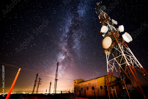 From below wonderful view of media antenna and silhouettes of electricity high voltage power towers against night city on horizon and gorgeous Milky Way in clear sky