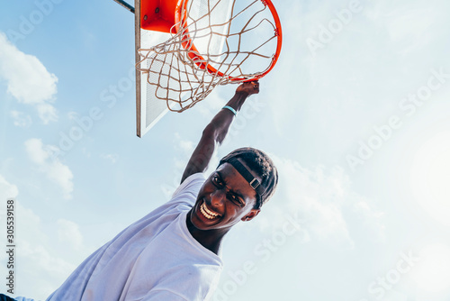 Powerful energetic African American sportsman hanging on basketball lap after scoring ball in net on playground