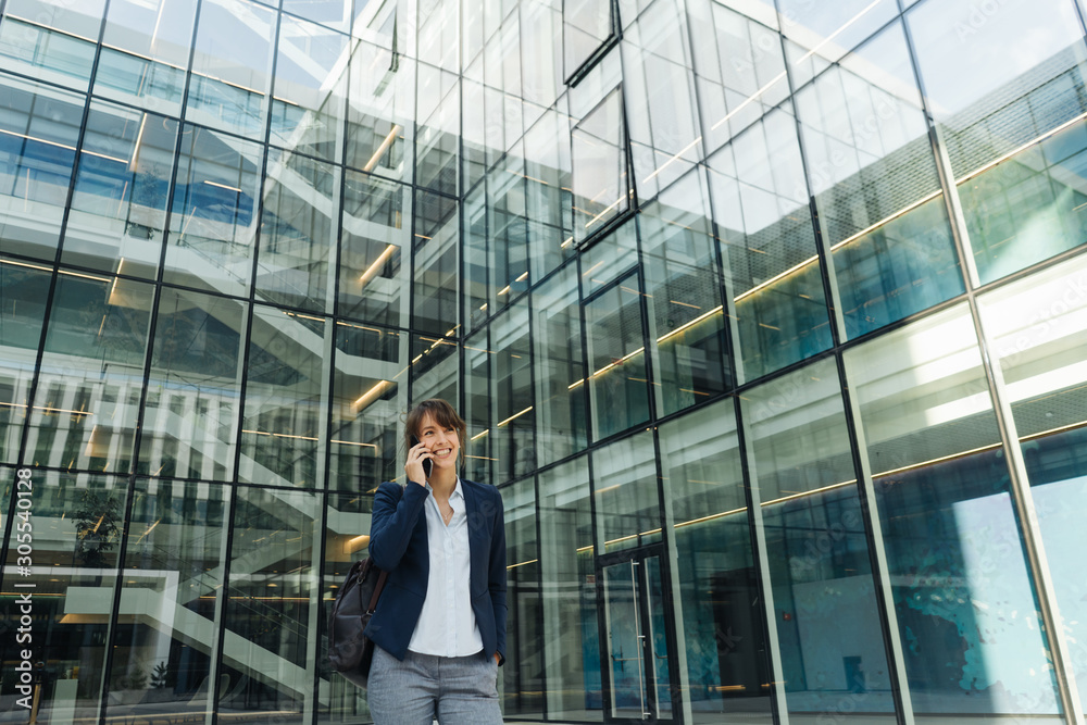 Low angle of happy businesswoman holding hand in pocket and using smartphone while standing outside business center on city street Low angle of happy businesswoman holding hand in pocket and using smartphone while standing outside business center on city street
