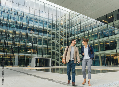 Full body delighted colleagues smiling and looking at each other while walking outside contemporary building after work