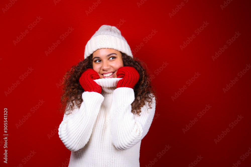 Studio portrait of young woman with dark skin and long curly hair wearing knitted turtle neck sweater over the festive red wall with a lot of copy space for text. Close up, isolated background. Studio portrait of young woman with dark skin and long curly hair wearing knitted turtle neck sweater over the festive red wall with a lot of copy space for text. Close up, isolated background.