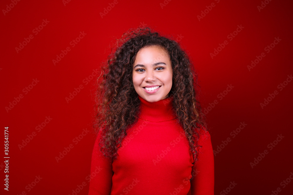 Studio portrait of young woman with dark skin and long curly hair wearing knitted turtle neck sweater over the festive red wall with a lot of copy space for text. Close up, isolated background. Studio portrait of young woman with dark skin and long curly hair wearing knitted turtle neck sweater over the festive red wall with a lot of copy space for text. Close up, isolated background.