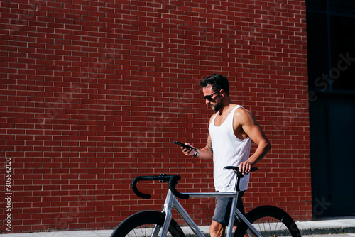 Side view of handsome male cyclist in sportswear and sunglasses using smartphone while standing with bike next to red brick wall photo