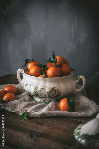 Sophisticated still life with ripe orange tangerines with green leaves in deep white bowl on table on gray background