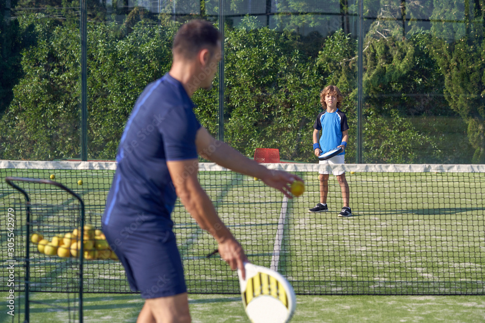 Boy in sportswear waiting for coach passing ball while playing paddle tennis on court during training