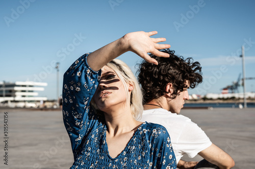 Attractive pensive woman covering self with hand from sunlight and through fingers while sitting back to back with young man and dreaming against blue sky and blurred modern buildings on square