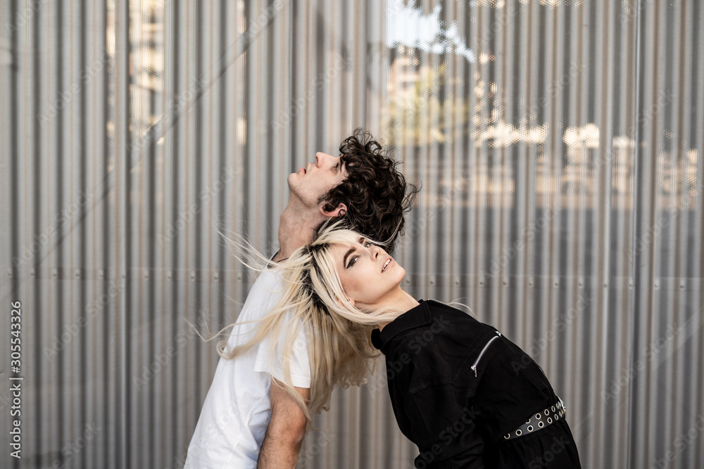 Side view of trendy couple supporting each other while standing back to back and dreaming against striped wall of modern building Side view of trendy couple supporting each other while standing back to back and dreaming against striped wall of modern building