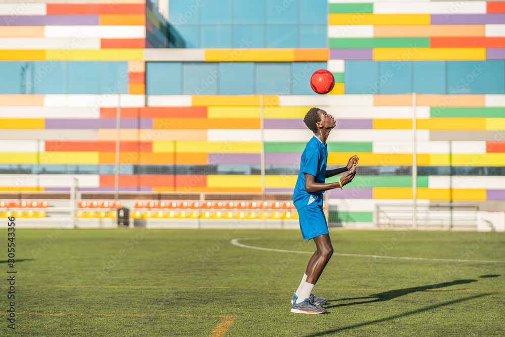 Full body side view of African American teenager in blue uniform bouncing bright ball on head during training on football field Full body side view of African American teenager in blue uniform bouncing bright ball on head during training on football field