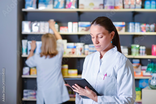 Beautiful young pharmacist standing next to medicine shelves, holding tablet