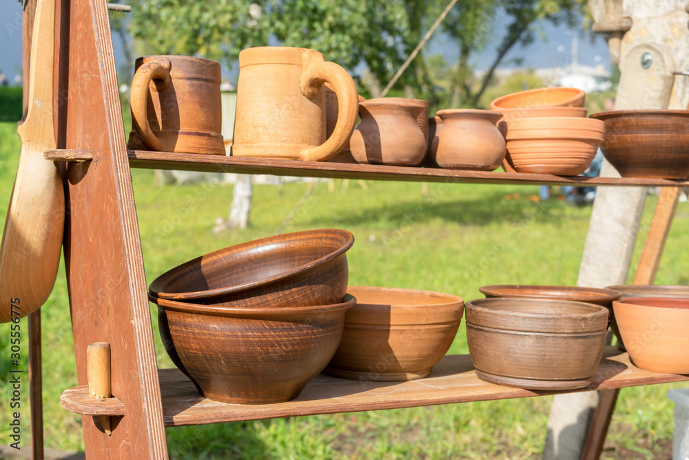 Many clay dishes on wooden shelf in Park. Pottery fair. Potter's craft. Kitchen utensils Many clay dishes on wooden shelf in Park. Pottery fair. Potter's craft. Kitchen utensils