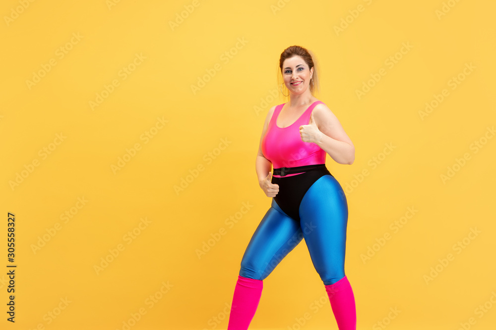 Young caucasian plus size female model's training on yellow background. Copyspace. Concept of sport, healthy lifestyle, body positive, fashion, style. Stylish woman posing with thumb up and smiling.