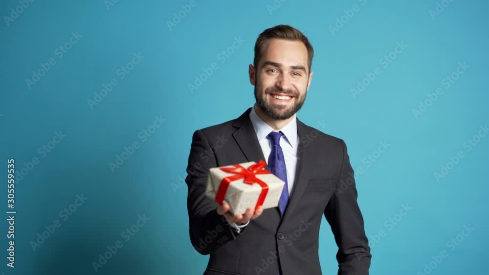 Handsome businessman in suit gives gift box and hands it to the camera. He is happy, smiling. Man with beard on blue background. Positive holiday footage