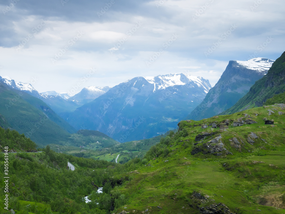 Mountain landscape with snow on the peaks near Geiranger, Norway and a beautiful green valley with a river and winding highway up to viewpoint on a cloudy day.
