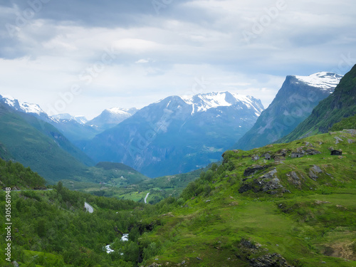 Mountain landscape with snow on the peaks near Geiranger, Norway and a beautiful green valley with a river and winding highway up to viewpoint on a cloudy day.