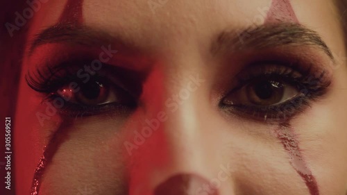 close-up portrait of a woman’s hazel eyes. clown makeup, bloody tears, long eyelashes, red night light, stripes on the face. Creative greasepaint art paints. creative fashion hororr design  photo