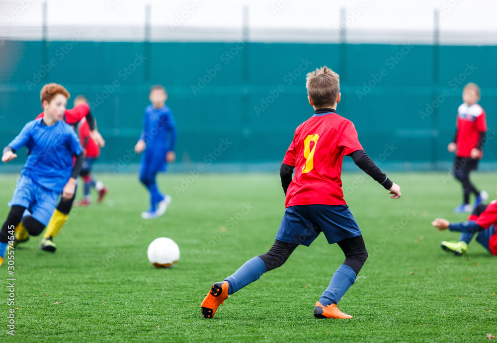 Boys in red and blue sportswear plays football on field, dribbles ball. Young soccer players with ball on green grass. Training, football, active lifestyle for kids concept 