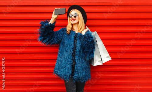 Attractive young smiling woman taking selfie picture by smartphone with shopping bags, stylish female model wearing blue faux fur coat, black round hat and sunglasses over red wall background © rohappy