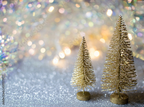 Two small gold Christmas tree on a silver surface with defocused background with bokeh lights. 