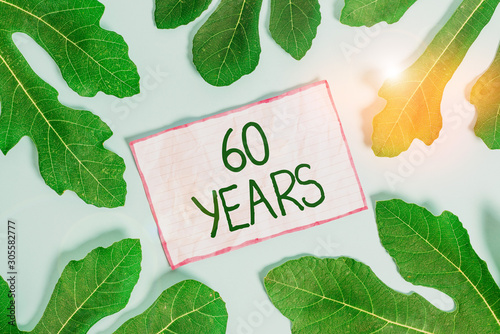 Handwriting text writing 60 Years. Conceptual photo It is the onset of a golden era Commemorating a special day Leaves surrounding notepaper above an empty soft pastel table as background