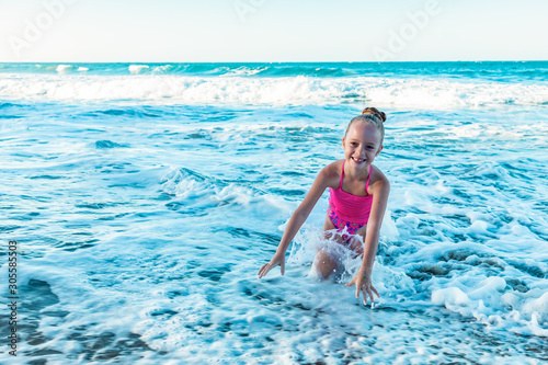 A playful cute little girl in a colorful pink swimsuit sitting on the sand, waves on the beach, ocean background, copy space