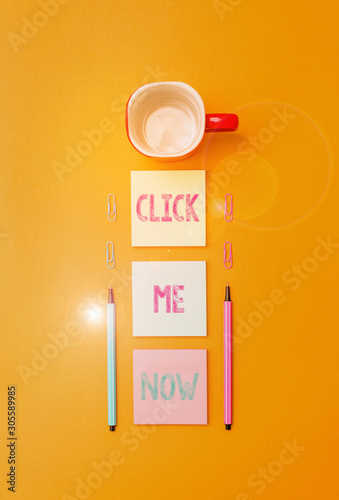 Text sign showing Click Me Now. Business photo text Internet helping desk Press the button Online Icon Nertwork Coffee cup blank colored sticky note two ballpoints clips yolk background photo