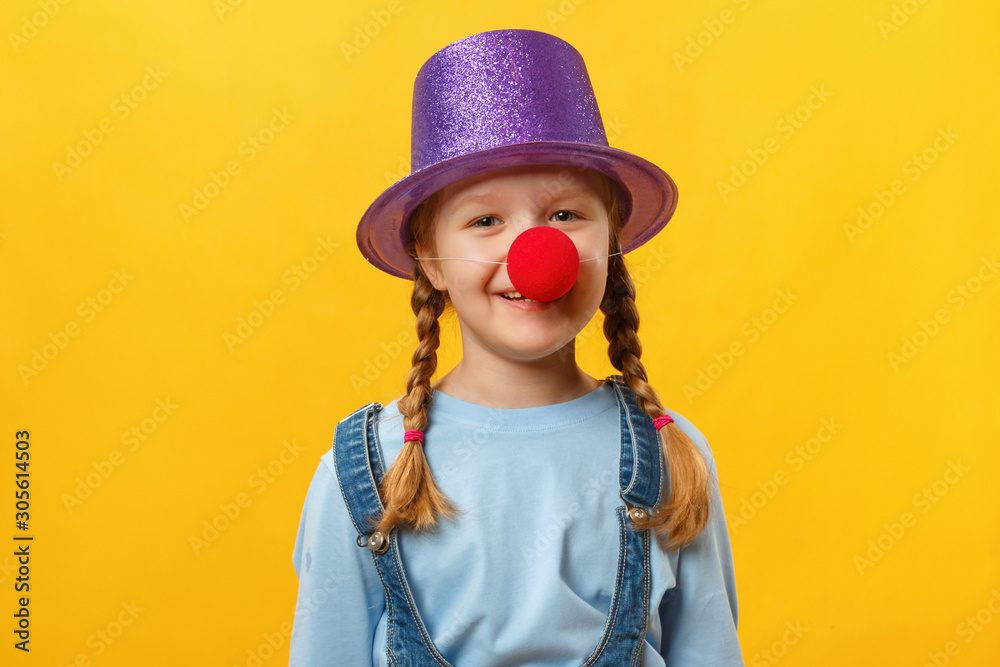 Funny child clown, hat and a red nose. Cheerful little girl on a yellow background. April 1. April Fools Day. Copy space