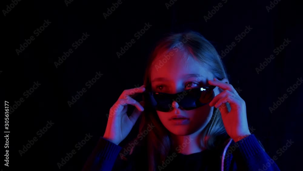 Slowmo teenager girl in black sunglasses looking around in dark room with red and blue light. Portrait cyberpunk girl chewing gum and wearing black glasses in dark studio with neon lighting