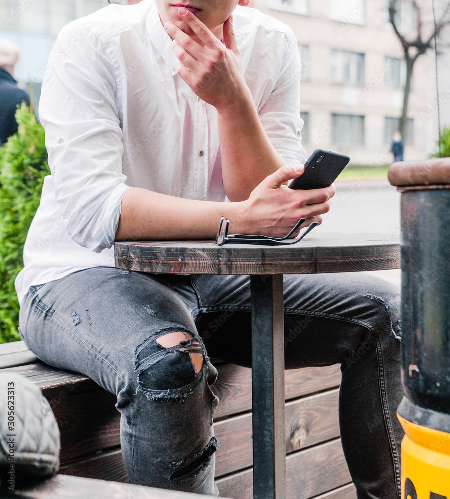 A young man in a white shirt and jeans sits at a table in a cafe on the street and looks at the phone