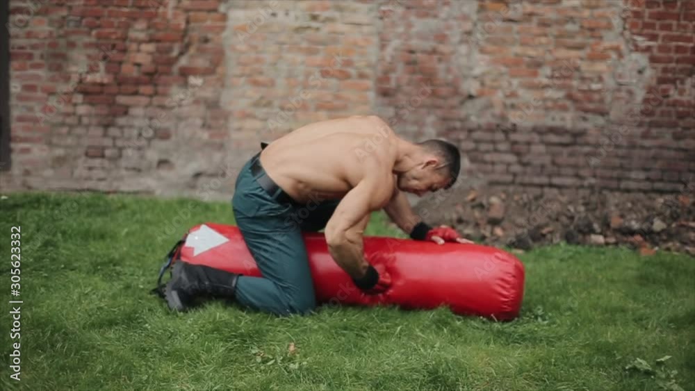 Muscular sportsman sitting on long punching bag outdoors, punching it, doing backflip, keep beating the bag Muscular sportsman sitting on long punching bag outdoors, punching it, doing backflip, keep beating the bag