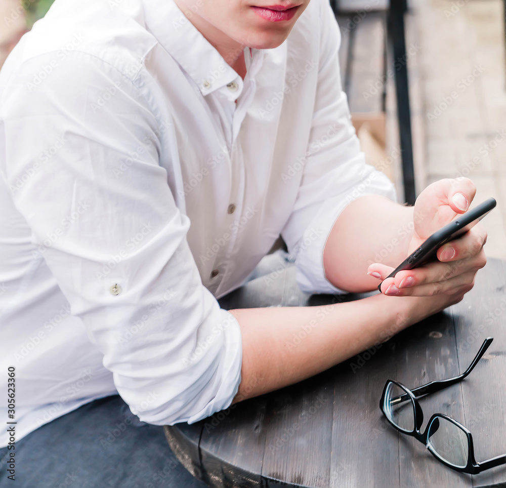 A young man in a white shirt and jeans sits at a table in a cafe, holds a phone in his hands and looks at the camera