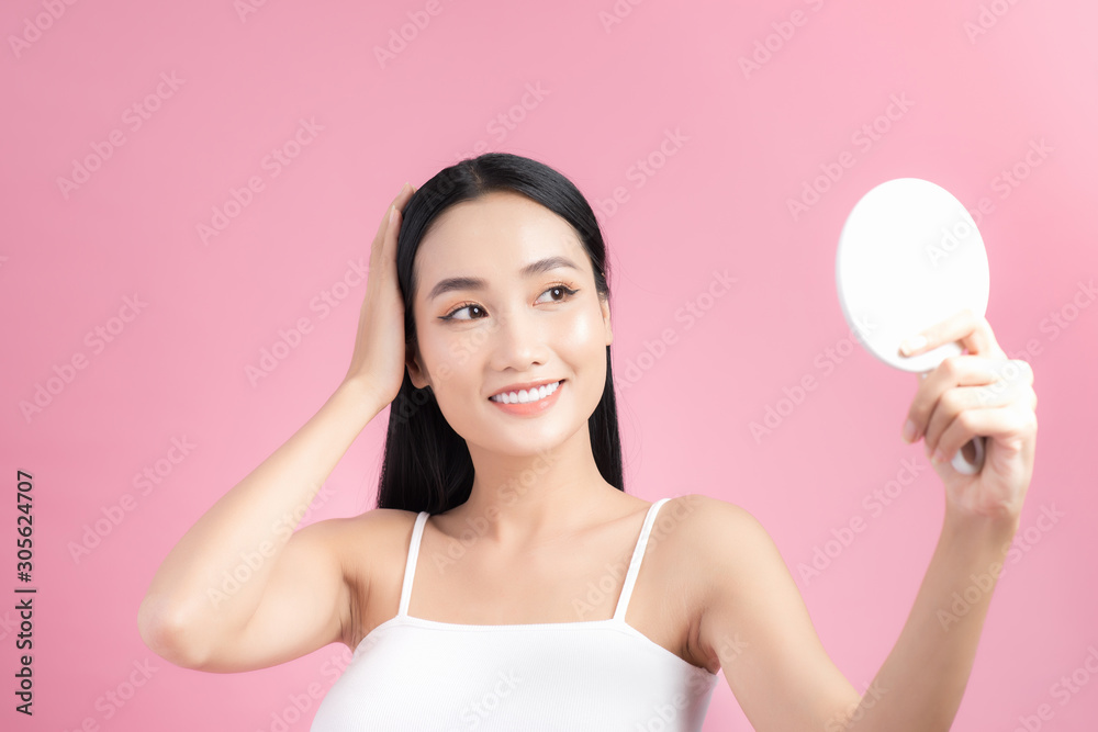 Beauty portrait of a cheerful attractive Asian woman examining her face while looking at the mirror isolated over pink background Beauty portrait of a cheerful attractive Asian woman examining her face while looking at the mirror isolated over pink background