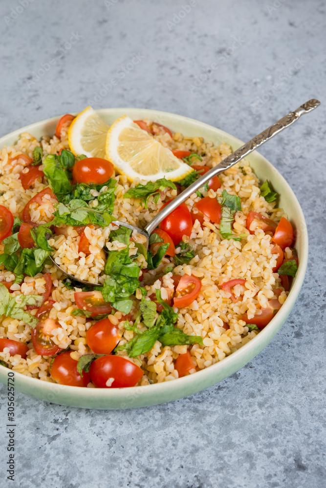 Tabouleh salad with tomatoes and fresh herbs in a plate with a spoon stands in the frame on a concrete table . Traditionally an Oriental dish. Tabouleh salad with tomatoes and fresh herbs in a plate with a spoon stands in the frame on a concrete table . Traditionally an Oriental dish.