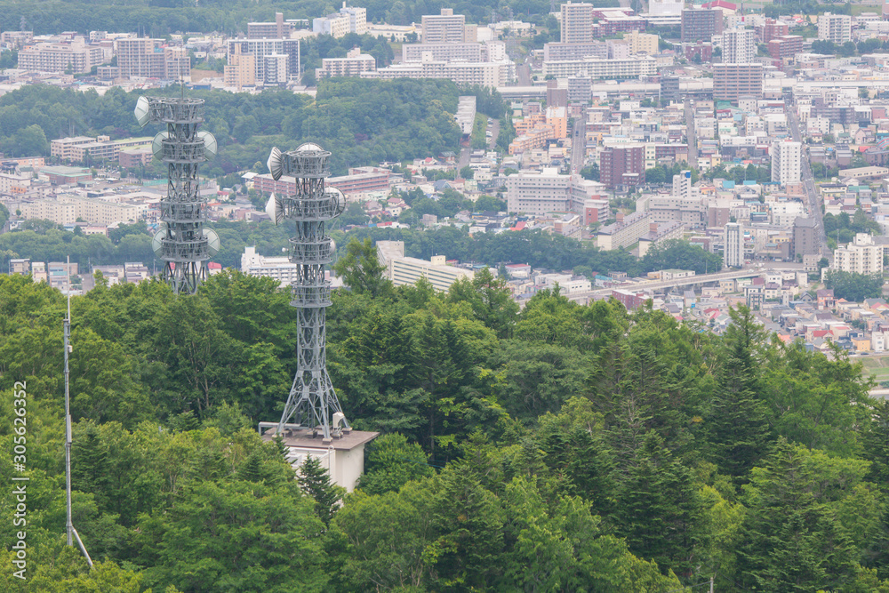 Telecommunication Signal Tower Antenna or Cellular Transmission Tower locate on hill surrounded with green trees and Sapporo City in background.