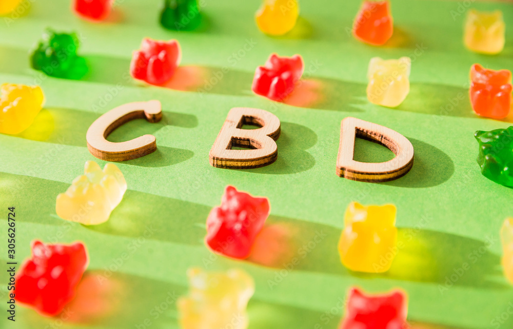 Naklejka premium Wooden letters CBD on a green background and gelatin in the form of bears. View from above. The shadow of the sun falls on the background