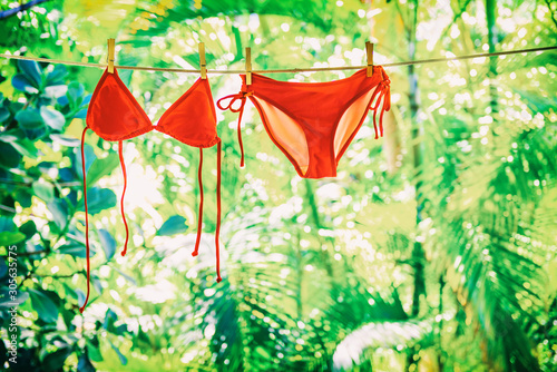 Clothing drying on clothesline outside on summer vacation handwash air dry eco friendly laundry. Red bikini swimming suit hanging outdoor women swimwear with green plants. photo