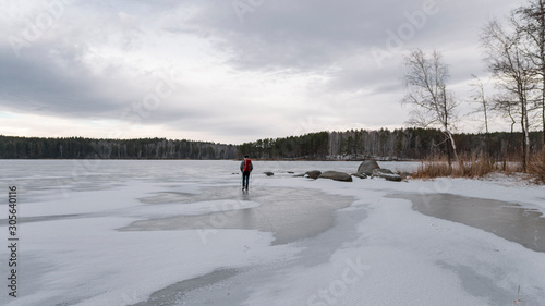 Young man walking on frozen lake with backpack and trekking poles; cloudy dramatic winter sky, cold weather, snowy lakesides with pine birch forest; abstract natural ice pattern; hiking lifestyle