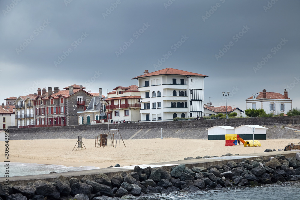Sandy beach, traditional houses and stone embankment of Saint-Jean-de-Luz, Basque Country, Atlantic coast, France. Coastal french town at cloudy sky summer day. Sea shore