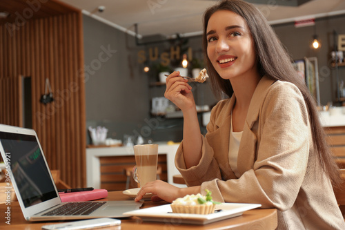 Young blogger with laptop eating cake in cafe