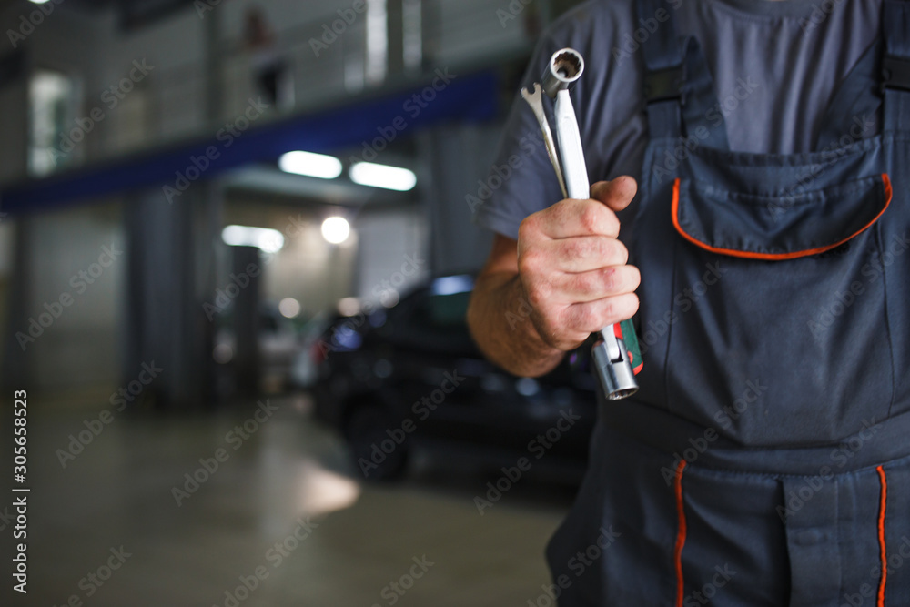 A mechanic in overalls with tools in his hands stands with his arms crossed on his chest. Work in the service station in the background. A mechanic in overalls with tools in his hands stands with his arms crossed on his chest. Work in the service station in the background.