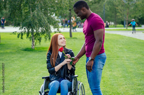 afro american man marriage proposal giving a ring to his redhaired ginger girlfriend.she sitting on wheel chair and surprised and amazed