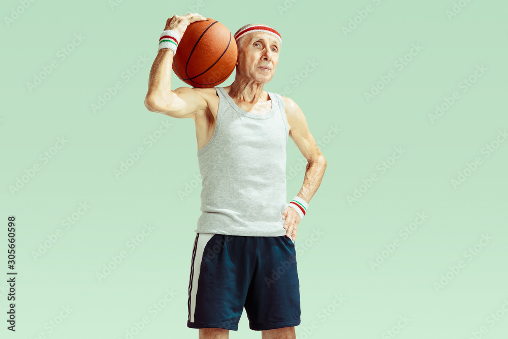 Senior man wearing sportwear playing basketball isolated on green background. Caucasian man in great shape stays active and sportive. Concept of sport, activity, movement, wellbeing. Copyspace, ad.