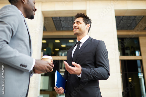 Young businessman in black suit smiling and explaining something to his African colleague while they meeting near the office building outdoors